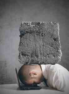 Image of a man with his head down on his laptop keyboard, crushed by a concrete block representing a budget. | The-Military-Guide.com