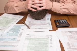 This image is surprisingly easy to find online. Or maybe not so surprisingly. Image of a man with his head down on his desk and his hands clenched over his head, in despair of how much he’ll have to pay on the income-tax forms scattered around him | MilitaryFinancialIndependence.com