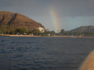 Image of Pokai Bay (Waianae, Oahu) from the middle of the bay (on a longboard) looking at the south shore beach park with a rainbow in front of the hill and the heiau. | MilitaryFinancialIndependence.com